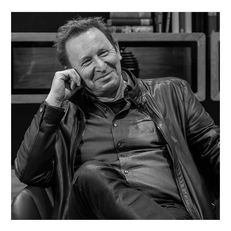 Martin Ballendat, de ontwerper van SHUFFLEis1, zit op een stoel, ondersteunt zijn hoofd met zijn rechterhand en glimlacht. Hij draagt een spijkerbroek, een overhemd en daaroverheen een leren jas.